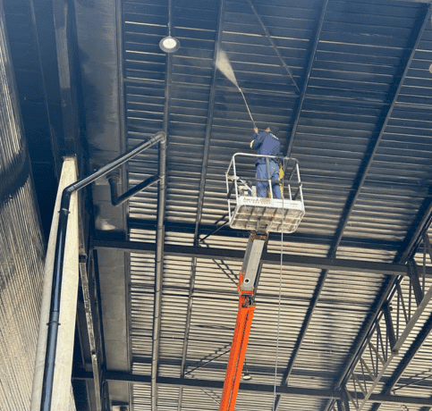 A worker on a raised platform power washes the interior ceiling of a large industrial building.